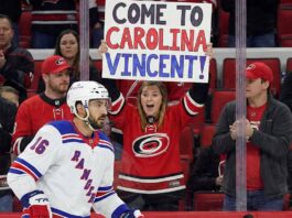 Is Vincent Trocheck Heading Home to the Hurricanes? Vincent Trocheck, wearing a New York Rangers jersey, skates on the ice during a game, while a female fan in a Carolina Hurricanes jersey behind the glass holds a sign that reads, "COME TO CAROLINA VINCENT!".