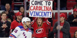Vincent Trocheck, wearing a New York Rangers jersey, skates on the ice during a game, while a female fan in a Carolina Hurricanes jersey behind the glass holds a sign that reads, "COME TO CAROLINA VINCENT!".