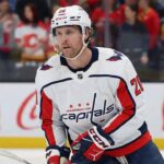 A Washington Capitals hockey player wearing a white away jersey with the number 20 and a white helmet, stands on the ice holding his stick during a game. He is looking forward with a focused expression. Fans wearing Calgary Flames jerseys are visible in the background.