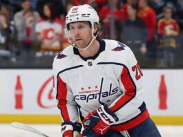 A Washington Capitals hockey player wearing a white away jersey with the number 20 and a white helmet, stands on the ice holding his stick during a game. He is looking forward with a focused expression. Fans wearing Calgary Flames jerseys are visible in the background.