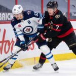 Winnipeg Jets center Mark Scheifele (white away jersey) and Ottawa Senators captain Brady Tkachuk (black home jersey) battle fiercely for the puck along the arena boards during an NHL game.