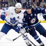 Toronto Maple Leafs center John Tavares (white jersey, 91) and Winnipeg Jets center Mark Scheifele (dark blue jersey, 55) battle stick-to-stick for control of the hockey puck during an NHL game.
