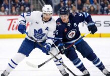 Toronto Maple Leafs center John Tavares (white jersey, 91) and Winnipeg Jets center Mark Scheifele (dark blue jersey, 55) battle stick-to-stick for control of the hockey puck during an NHL game.