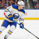 Is Zach Whitecloud the Missing Piece for the Buffalo Sabres’ Playoff Run? Buffalo Sabres defenseman Zach Whitecloud, wearing number 2 in a white away jersey with blue and gold trim, skates with the puck across the ice during a game. Fans and rink boards are visible in the background.