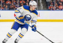 Buffalo Sabres defenseman Zach Whitecloud, wearing number 2 in a white away jersey with blue and gold trim, skates with the puck across the ice during a game. Fans and rink boards are visible in the background.