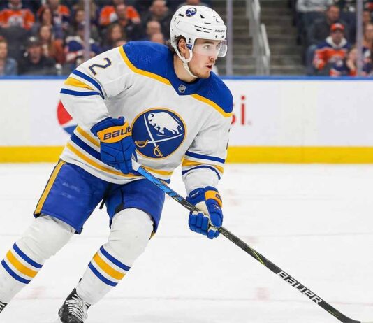 Is Zach Whitecloud the Missing Piece for the Buffalo Sabres’ Playoff Run? Buffalo Sabres defenseman Zach Whitecloud, wearing number 2 in a white away jersey with blue and gold trim, skates with the puck across the ice during a game. Fans and rink boards are visible in the background.