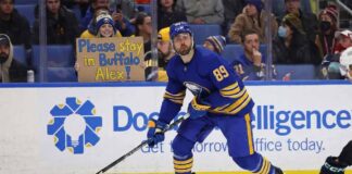 Buffalo Sabres forward Alex Tuch, wearing number 89 in the blue home jersey, skates with the puck during a game. Behind him, in the spectator stands, a female fan wearing a Sabres winter hat holds up a cardboard sign with blue and yellow lettering that reads, "Please stay in Buffalo Alex!".