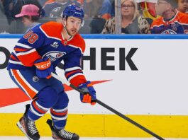 An Edmonton Oilers player, wearing number 88 on his blue and orange jersey, skates with the puck along the boards during a hockey game. He is looking forward, and spectators are visible in the background behind the glass.