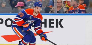 An Edmonton Oilers player, wearing number 88 on his blue and orange jersey, skates with the puck along the boards during a hockey game. He is looking forward, and spectators are visible in the background behind the glass.