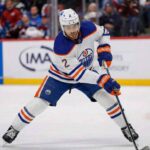 Artem Zub, wearing a white Edmonton Oilers away jersey with the number 2, is pictured on the ice in a low stance, handling a hockey puck with his stick.