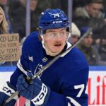 Toronto Maple Leafs player Bobby McMann (74) skates on the ice while a fan behind the glass to the left holds a handwritten cardboard sign that reads, "GOOD LUCK WITH YOUR NEW TEAM BOBBY!".
