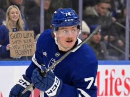 Toronto Maple Leafs player Bobby McMann (74) skates on the ice while a fan behind the glass to the left holds a handwritten cardboard sign that reads, "GOOD LUCK WITH YOUR NEW TEAM BOBBY!".
