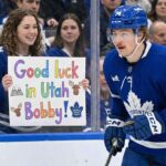 Will the Toronto Maple Leafs Trade Bobby McMann to Utah? A photograph taken from behind the glass shows Toronto Maple Leafs forward Bobby McMann (number 74, wearing a blue helmet and jersey) in motion on the ice. Behind the clear barrier, a smiling young female fan wearing a Leafs jersey holds up a colorful hand-drawn sign. The sign features drawings of mountains and moose and prominently reads: "Good luck in Utah Bobby!". Other spectators wearing Leafs gear fill the background crowd.