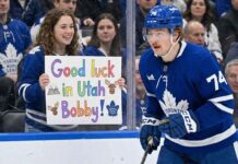 A photograph taken from behind the glass shows Toronto Maple Leafs forward Bobby McMann (number 74, wearing a blue helmet and jersey) in motion on the ice. Behind the clear barrier, a smiling young female fan wearing a Leafs jersey holds up a colorful hand-drawn sign. The sign features drawings of mountains and moose and prominently reads: "Good luck in Utah Bobby!". Other spectators wearing Leafs gear fill the background crowd.