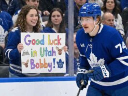 Will the Toronto Maple Leafs Trade Bobby McMann to Utah? A photograph taken from behind the glass shows Toronto Maple Leafs forward Bobby McMann (number 74, wearing a blue helmet and jersey) in motion on the ice. Behind the clear barrier, a smiling young female fan wearing a Leafs jersey holds up a colorful hand-drawn sign. The sign features drawings of mountains and moose and prominently reads: "Good luck in Utah Bobby!". Other spectators wearing Leafs gear fill the background crowd.