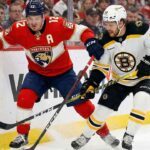 A close-up photograph of Florida Panthers forward Matthew Tkachuk (12), wearing a red home jersey with an 'A', and Boston Bruins David Pastrnak (88), in a white away jersey, battling with their sticks for a loose puck along the rink boards during an NHL game. Ice spray is visible around their skates as they jostle for position.