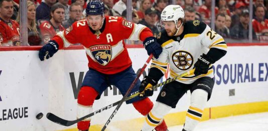 A close-up photograph of Florida Panthers forward Matthew Tkachuk (12), wearing a red home jersey with an 'A', and Boston Bruins David Pastrnak (88), in a white away jersey, battling with their sticks for a loose puck along the rink boards during an NHL game. Ice spray is visible around their skates as they jostle for position.