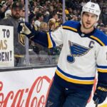 St. Louis Blues captain Brayden Schenn, wearing his white away jersey with the 'C' patch, skates along the boards at T-Mobile Arena, looking towards a female fan behind the glass holding a handmade sign that reads "COME TO VEGAS BRAYDEN!".