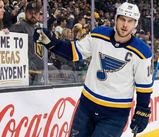 St. Louis Blues captain Brayden Schenn, wearing his white away jersey with the 'C' patch, skates along the boards at T-Mobile Arena, looking towards a female fan behind the glass holding a handmade sign that reads "COME TO VEGAS BRAYDEN!".