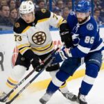 Boston Bruins defenseman Charlie McAvoy (73) in a white jersey battles for puck possession against Tampa Bay Lightning forward Nikita Kucherov (86) in a blue jersey along the boards during an NHL game. Their sticks are clashing near the puck as fans watch in the background.