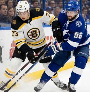 Boston Bruins defenseman Charlie McAvoy (73) in a white jersey battles for puck possession against Tampa Bay Lightning forward Nikita Kucherov (86) in a blue jersey along the boards during an NHL game. Their sticks are clashing near the puck as fans watch in the background.