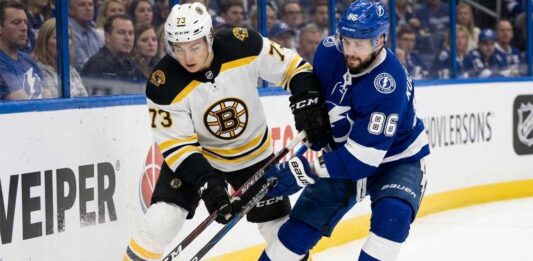 Boston Bruins defenseman Charlie McAvoy (73) in a white jersey battles for puck possession against Tampa Bay Lightning forward Nikita Kucherov (86) in a blue jersey along the boards during an NHL game. Their sticks are clashing near the puck as fans watch in the background.