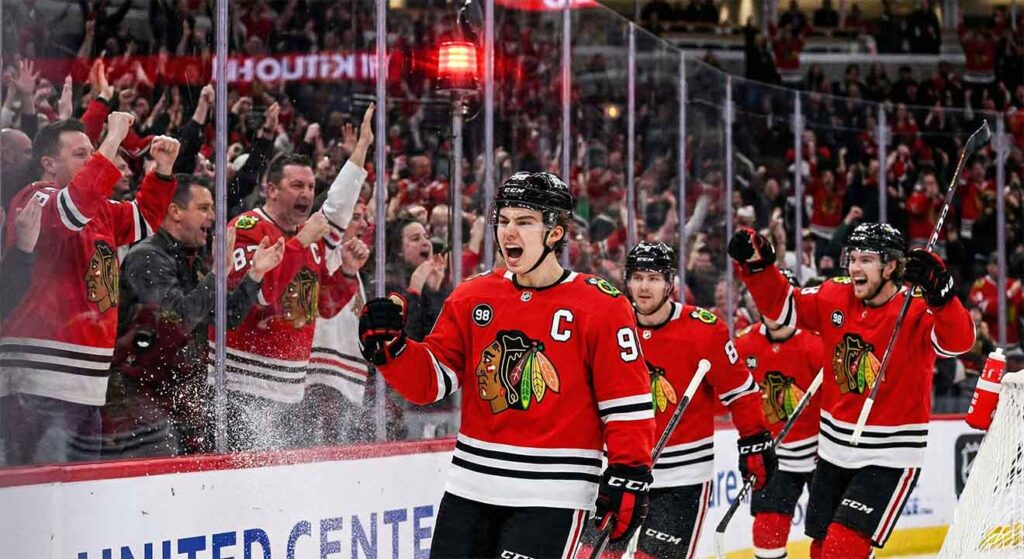 Chicago Blackhawks captain Connor Bedard, wearing his number 98 jersey, screams in celebration and pumps his fist after scoring a goal, as teammates join him on the ice and fans cheer wildly behind the glass with the red goal light illuminated.