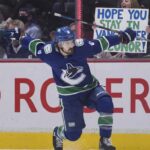 Vancouver Canucks forward Conor Garland (#8) celebrates a goal by sliding on one knee across the ice with his arms extended. Behind the glass boards featuring the Rogers logo, cheering fans are visible, including a woman holding up a handmade white sign with blue and green lettering that reads, "HOPE YOU STAY IN VANCOUVER CONOR!".