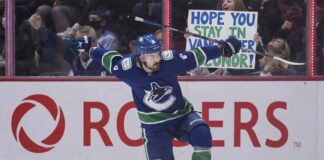 Vancouver Canucks forward Conor Garland (#8) celebrates a goal by sliding on one knee across the ice with his arms extended. Behind the glass boards featuring the Rogers logo, cheering fans are visible, including a woman holding up a handmade white sign with blue and green lettering that reads, "HOPE YOU STAY IN VANCOUVER CONOR!".