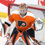 Philadelphia Flyers goaltender Dennis Hildeby is pictured in a crouched, ready stance in front of the net, wearing the team's orange, black, and white home jersey, pads, and a custom-painted mask. A hockey puck is mid-air, just in front of his leg pads.