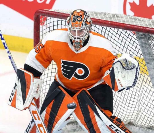 Philadelphia Flyers goaltender Dennis Hildeby is pictured in a crouched, ready stance in front of the net, wearing the team's orange, black, and white home jersey, pads, and a custom-painted mask. A hockey puck is mid-air, just in front of his leg pads.