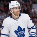 A photorealistic rendering of defenseman Dougie Hamilton standing on an ice hockey rink, wearing a white Toronto Maple Leafs away jersey with the blue Maple Leaf crest, blue gloves, and a white helmet bearing his number 7 and team decals.