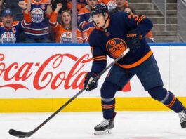 Chicago Blackhawks defenseman Connor Murphy depicted wearing an Edmonton Oilers dark blue home uniform, skating with a hockey stick on the ice in front of a red and white Coca-Cola advertisement on the arena boards.