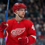 Elias Pettersson, wearing a Detroit Red Wings jersey and helmet, looks towards the right side of the frame while holding a hockey stick on the ice.