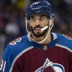 Close-up photograph of NHL forward Evander Kane wearing a burgundy and blue Colorado Avalanche home jersey with number 91 on the sleeve and helmet. He is looking off-camera to the left with a white mouthguard dangling from his mouth against a blurred arena crowd background.