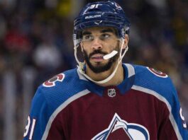 Close-up photograph of NHL forward Evander Kane wearing a burgundy and blue Colorado Avalanche home jersey with number 91 on the sleeve and helmet. He is looking off-camera to the left with a white mouthguard dangling from his mouth against a blurred arena crowd background.