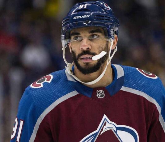 Close-up photograph of NHL forward Evander Kane wearing a burgundy and blue Colorado Avalanche home jersey with number 91 on the sleeve and helmet. He is looking off-camera to the left with a white mouthguard dangling from his mouth against a blurred arena crowd background.