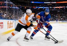 An action photograph from an NHL game showing Philadelphia Flyers forward Travis Konecny (left, in white #11 jersey) and New York Rangers center Mika Zibanejad (right, in blue #93 jersey) intensely battling for control of a loose puck on the ice.