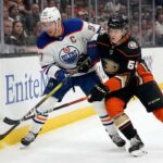 Edmonton Oilers captain Connor McDavid (#97) in a white away jersey and Anaheim Ducks forward Cutter Gauthier (#64) in a black home jersey battle physically for puck possession against the rink boards during an NHL game.
