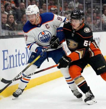 You Won’t Believe Who’s Out: Edmonton Oilers at Anaheim Ducks TV Guide & Prediction Edmonton Oilers captain Connor McDavid (#97) in a white away jersey and Anaheim Ducks forward Cutter Gauthier (#64) in a black home jersey battle physically for puck possession against the rink boards during an NHL game.