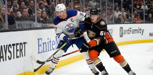 Edmonton Oilers captain Connor McDavid (#97) in a white away jersey and Anaheim Ducks forward Cutter Gauthier (#64) in a black home jersey battle physically for puck possession against the rink boards during an NHL game.