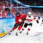 Will Team Canada Crush Team Switzerland Again? How to Watch Live! A Team Canada player in a red jersey battles for the puck against a Team Switzerland player in white near the arena boards during a women's ice hockey game at the Milano Cortina 2026 Winter Olympics. Fans wave Canadian flags in the background and Olympic branding is visible on the ice and boards.