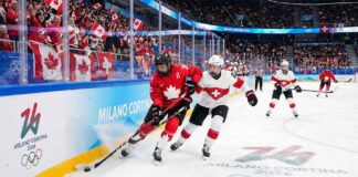 A Team Canada player in a red jersey battles for the puck against a Team Switzerland player in white near the arena boards during a women's ice hockey game at the Milano Cortina 2026 Winter Olympics. Fans wave Canadian flags in the background and Olympic branding is visible on the ice and boards.