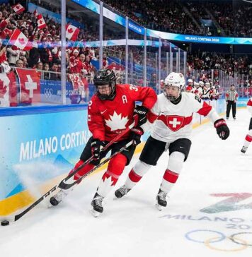 Will Team Canada Crush Team Switzerland Again? How to Watch Live! A Team Canada player in a red jersey battles for the puck against a Team Switzerland player in white near the arena boards during a women's ice hockey game at the Milano Cortina 2026 Winter Olympics. Fans wave Canadian flags in the background and Olympic branding is visible on the ice and boards.