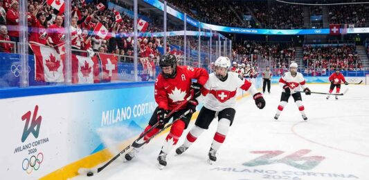 A Team Canada player in a red jersey battles for the puck against a Team Switzerland player in white near the arena boards during a women's ice hockey game at the Milano Cortina 2026 Winter Olympics. Fans wave Canadian flags in the background and Olympic branding is visible on the ice and boards.