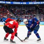 Marie-Philip Poulin in a red Team Canada jersey and Hilary Knight in a blue Team USA jersey crouch for a face-off during the women's ice hockey gold medal game at the 2026 Winter Olympics, with a packed crowd and scoreboard visible in the background indicating the event.
