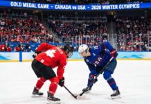 Marie-Philip Poulin in a red Team Canada jersey and Hilary Knight in a blue Team USA jersey crouch for a face-off during the women's ice hockey gold medal game at the 2026 Winter Olympics, with a packed crowd and scoreboard visible in the background indicating the event.