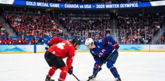 Marie-Philip Poulin in a red Team Canada jersey and Hilary Knight in a blue Team USA jersey crouch for a face-off during the women's ice hockey gold medal game at the 2026 Winter Olympics, with a packed crowd and scoreboard visible in the background indicating the event.