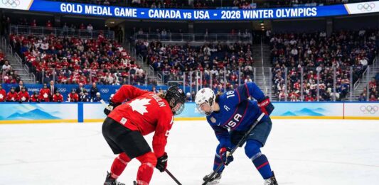 Marie-Philip Poulin in a red Team Canada jersey and Hilary Knight in a blue Team USA jersey crouch for a face-off during the women's ice hockey gold medal game at the 2026 Winter Olympics, with a packed crowd and scoreboard visible in the background indicating the event.
