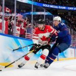 Team Canada captain Connor McDavid battles intensely with Team USA defenseman Charlie McAvoy for a loose puck along the arena boards during a men's Olympic gold medal ice hockey game. Fans wave Canadian and American flags in the background.
